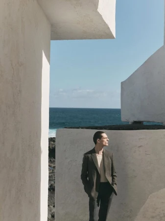 Man in blazer and trousers posing against textured white walls with a sea and sky background in a fashion editorial photo.