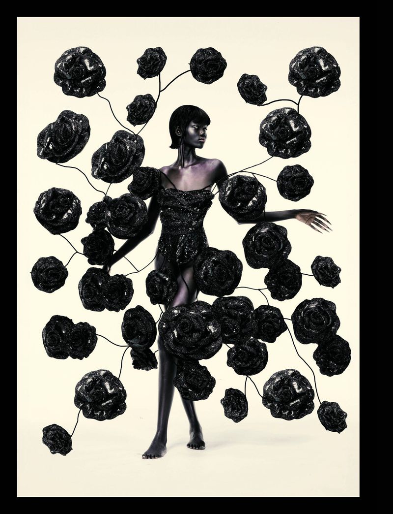 A dark-skinned woman in a sparkly black dress stands on a white floor surrounded by 3D-rendered black roses in a studio setting.