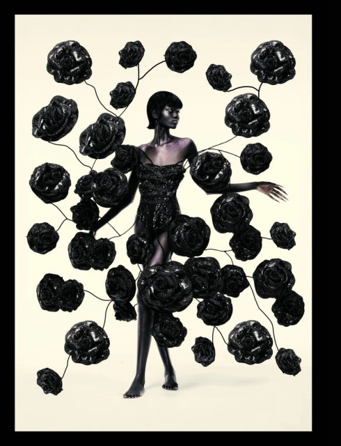 A dark-skinned woman in a sparkly black dress stands on a white floor surrounded by 3D-rendered black roses in a studio setting.