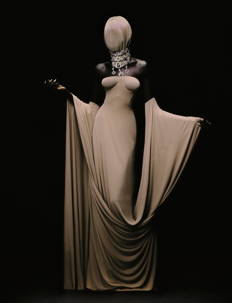 A model wears a draped beige gown with a head covering and elaborate crystal necklace against a dark background in a studio shot.