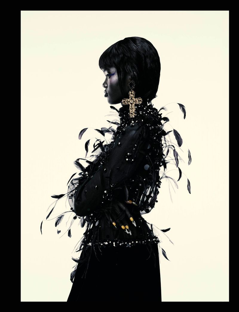 Profile shot of a Black woman wearing a black feather and beaded top, and a large cross earring against a neutral backdrop.