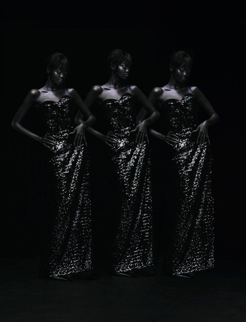 Three women with dark skin, wearing strapless sparkling dresses, pose in a dark studio with hands on hips, full shot.