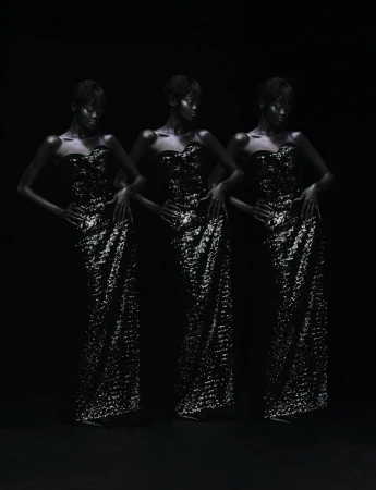 Three women with dark skin, wearing strapless sparkling dresses, pose in a dark studio with hands on hips, full shot.