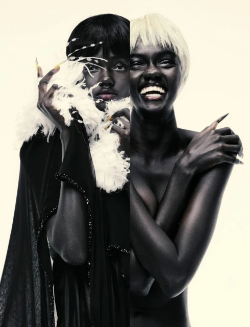 Striking fashion portrait featuring two women with dramatic makeup, wigs, and costumes against a cream background in a studio setting.