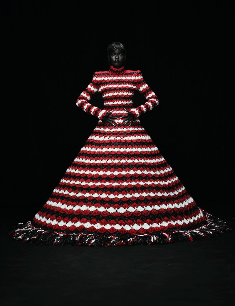 A black female fashion model poses in a large, red, white, and black gown with a dark background in a studio fashion shot.