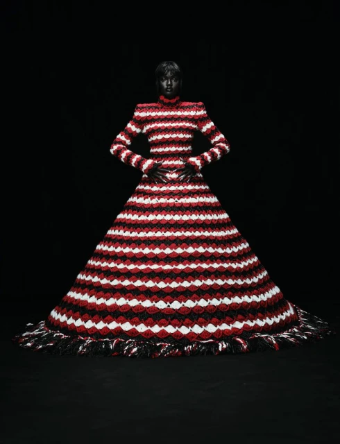A black female fashion model poses in a large, red, white, and black gown with a dark background in a studio fashion shot.