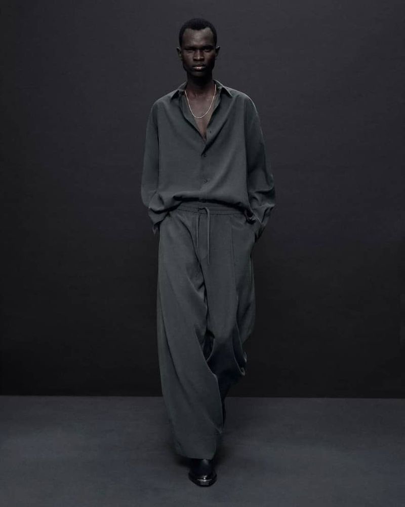 An adult male of African descent poses in a long-sleeved shirt and loose-fitting trousers against a dark grey backdrop indoors.