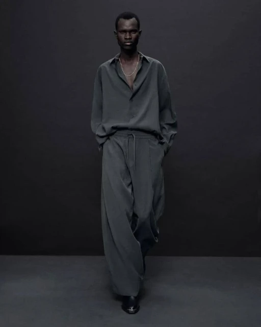 An adult male of African descent poses in a long-sleeved shirt and loose-fitting trousers against a dark grey backdrop indoors.
