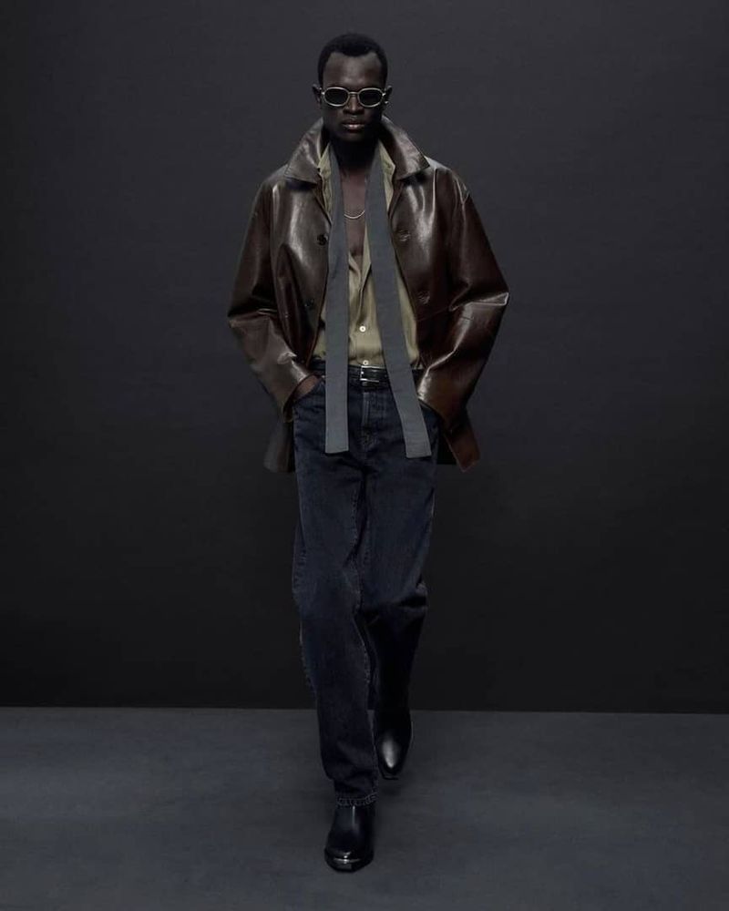 Man in a brown leather jacket, blue jeans, and black boots is walking in a photo studio setting. He is wearing sunglasses.