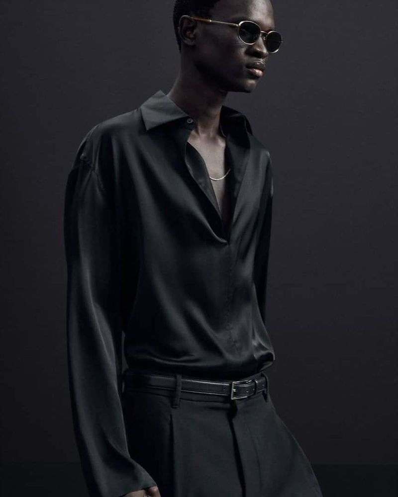Dark-skinned adult male poses in a black long-sleeve silk shirt and dark pants against a dark backdrop, showcasing a minimalist fashion aesthetic.