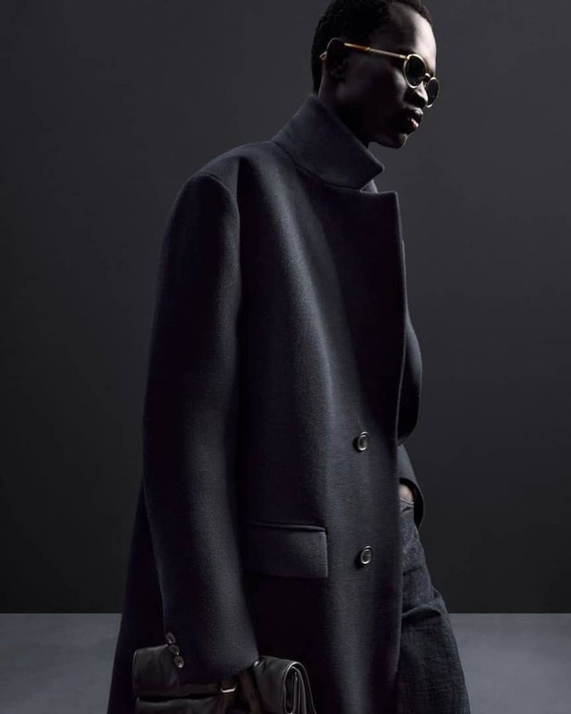 Dark-skinned man in a dark coat and round glasses holding a dark bag, posing indoors against a gray background in a studio-style shot.