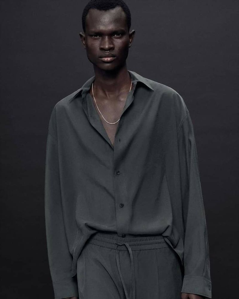 A dark-skinned male model poses in a relaxed button-down shirt and pants. The model is centered against a dark gray backdrop.