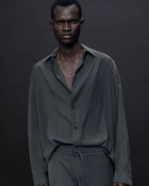 A dark-skinned male model poses in a relaxed button-down shirt and pants. The model is centered against a dark gray backdrop.