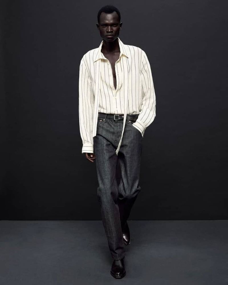 An adult male is shown standing against a grey background, wearing a vertically striped shirt, gray denim jeans and black dress shoes.