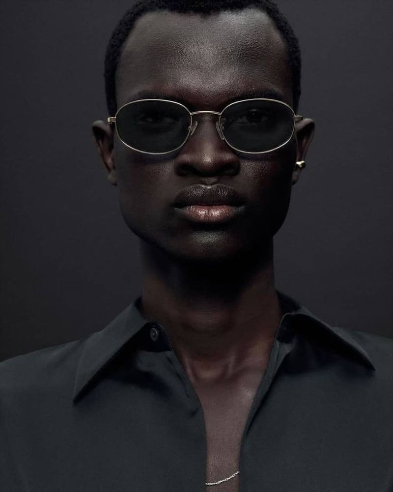 Portrait of a Black male model wearing sunglasses and a black collared shirt against a dark grey background in a studio setting.