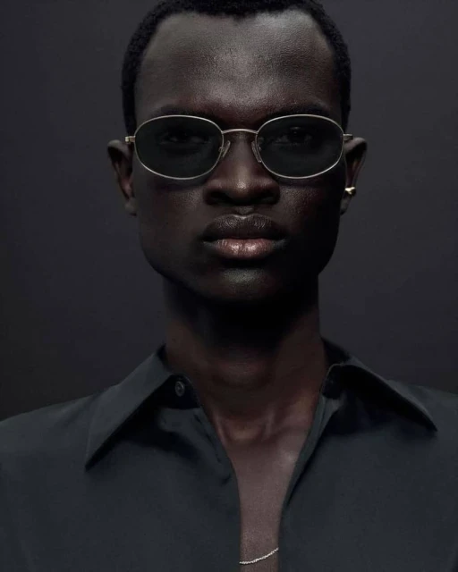 Portrait of a Black male model wearing sunglasses and a black collared shirt against a dark grey background in a studio setting.