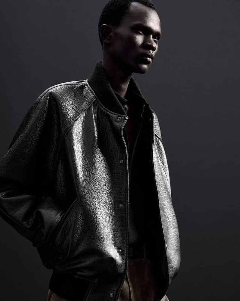 Fashion shot of an African-descent male model wearing a black leather-textured bomber jacket against a dark grey studio backdrop.
