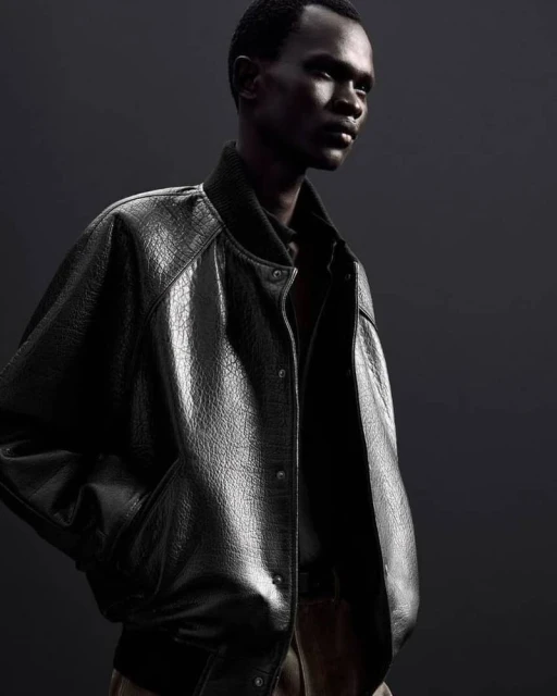 Fashion shot of an African-descent male model wearing a black leather-textured bomber jacket against a dark grey studio backdrop.