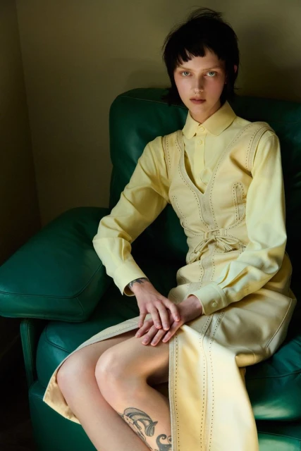 Adult female seated on a green leather couch wearing a yellow dress in a studio with soft lighting and neutral color palette.