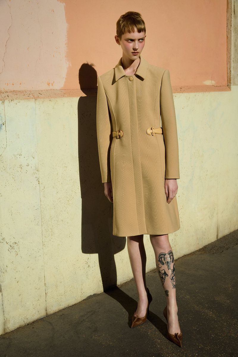 Fashionable woman in a tan coat standing outdoors with a leg tattoo and high heels in an urban setting with a textured wall.