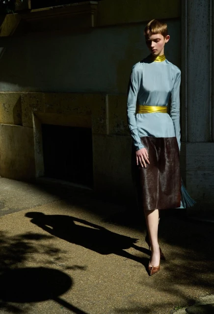 Woman poses in a long-sleeved, light blue top, brown skirt, and heels, standing on a sidewalk in front of a concrete wall.