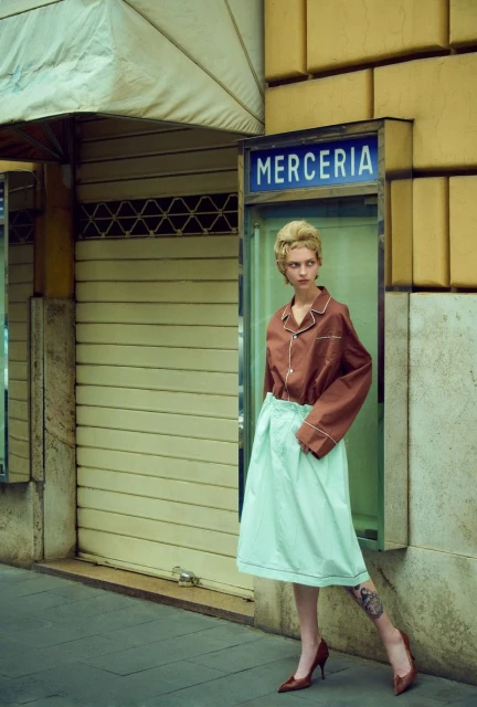 Adult female fashion model poses in front of a store wearing a brown top and light green skirt and brown heels. Outdoor shot.