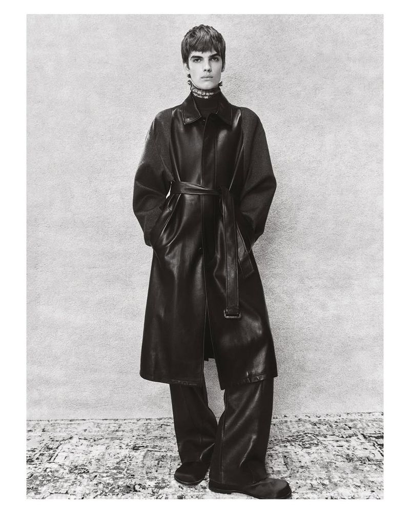 A person poses wearing a long black leather coat with a belt and matching black pants against a textured neutral backdrop.