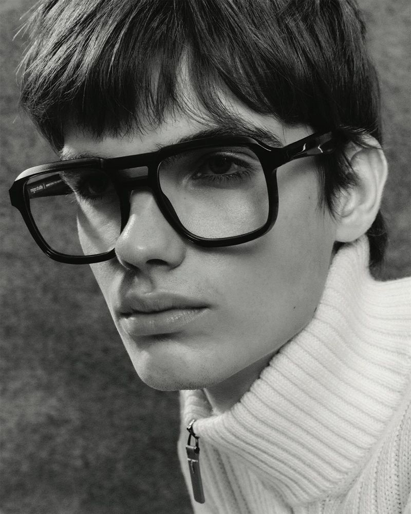 Close-up of a young adult man wearing black-rimmed glasses and a white turtleneck sweater against a grey studio backdrop.