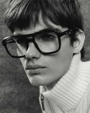 Close-up of a young adult man wearing black-rimmed glasses and a white turtleneck sweater against a grey studio backdrop.