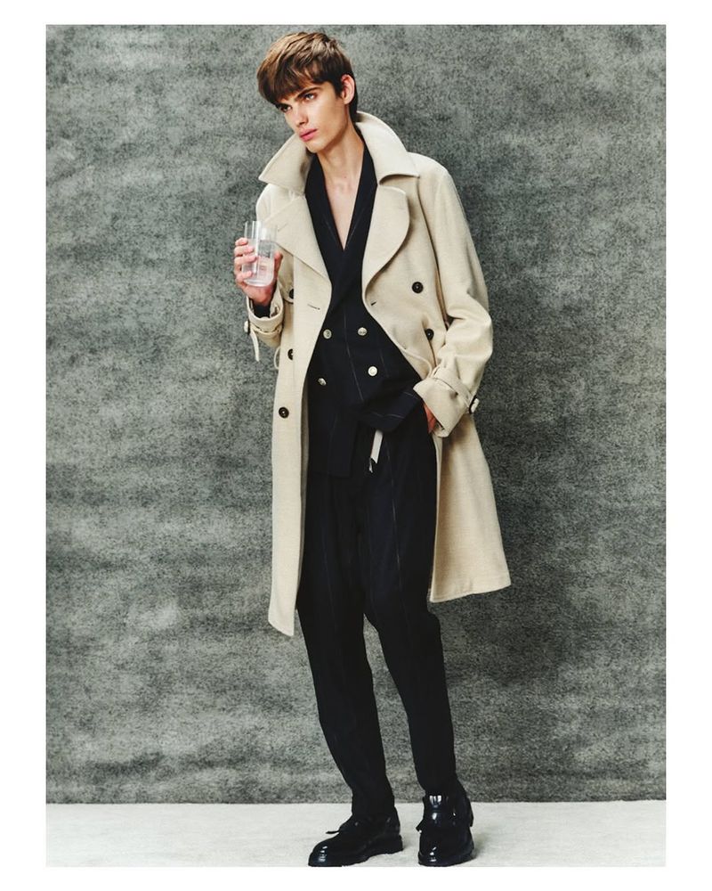 Male model in a beige trench coat and black suit poses against a textured gray background while holding a glass of water.