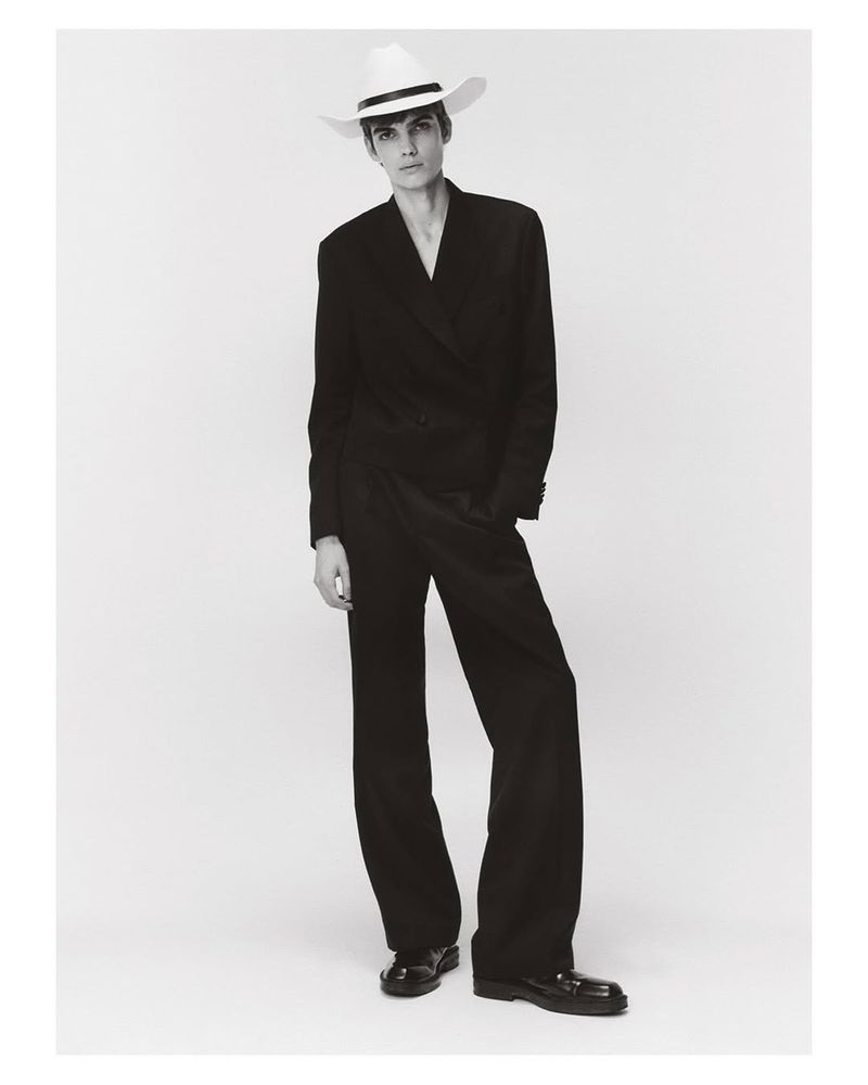 Monochrome portrait of a fashionable adult model in a dark suit and white hat, standing on a light background in a studio.