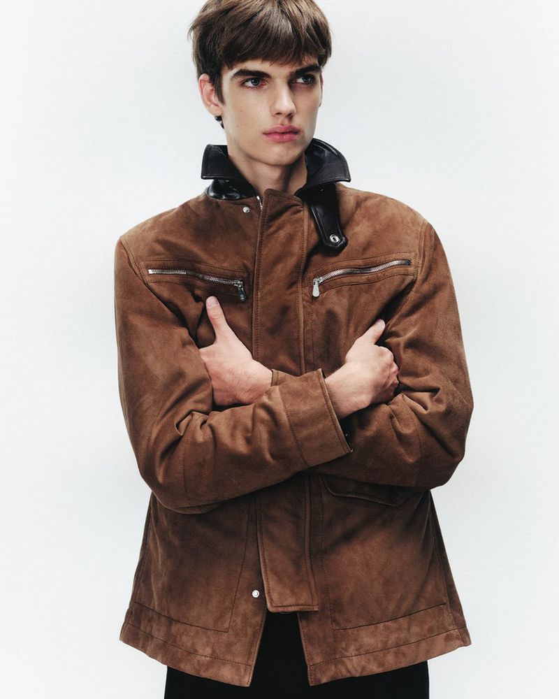Adult male poses in a brown jacket with a black collar against a white background, creating a simple yet fashionable portrait.