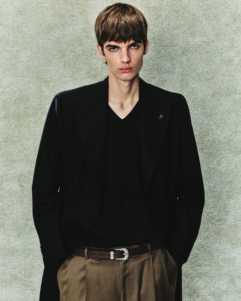 Fashion model in a black blazer, v-neck shirt, and pleated trousers with a western buckle belt against a textured grey backdrop.