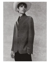 A male model wearing a light wide-brim cowboy hat and textured grey buttoned jacket posing against a mottled studio backdrop.