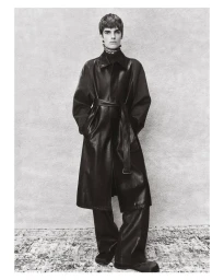 Male model wearing an oversized belted dark leather coat standing against a textured backdrop in a black and white fashion portrait.