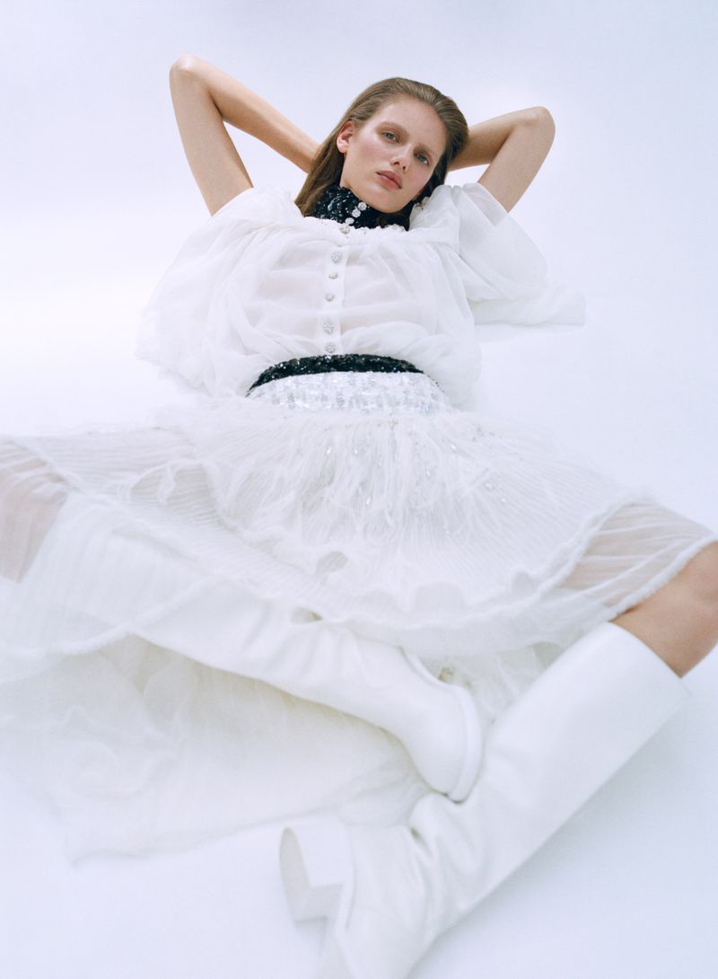A woman poses in a white outfit with white boots against a bright white backdrop in a fashion shot.