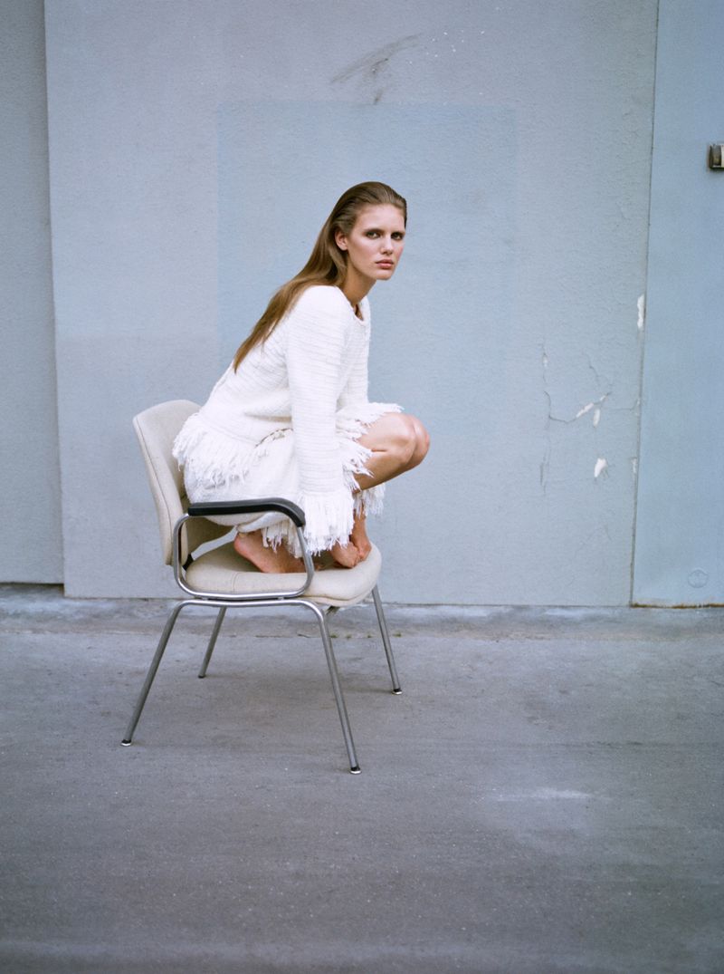 Woman in a white fringe dress poses on a chair against a gray wall, creating a minimalist fashion shot with a cool, editorial vibe.
