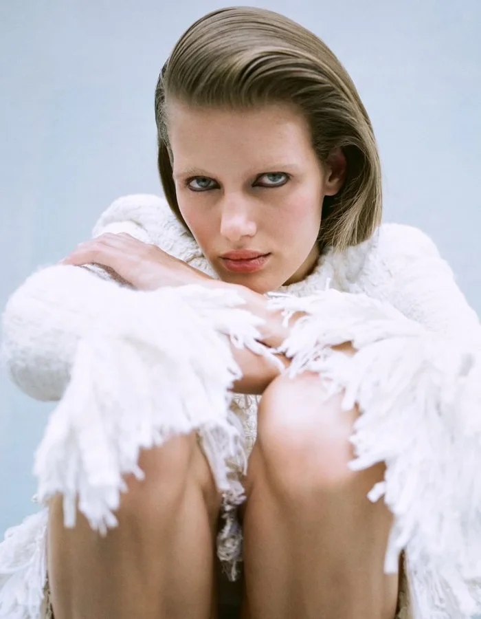 Model Rosalieke Fuchs seated in a white fringe knit sweater looking directly at camera against a pale blue seamless backdrop.