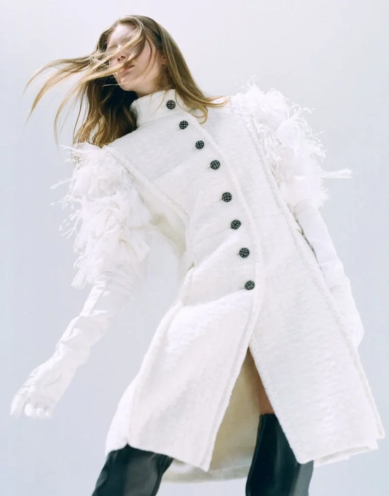 Model wearing a structured white buttoned coat with feathered shoulder trim and white opera gloves against a white studio background.