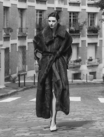 Woman in a long fur coat walks on a cobblestone street in front of an apartment building in a black and white fashion shot.