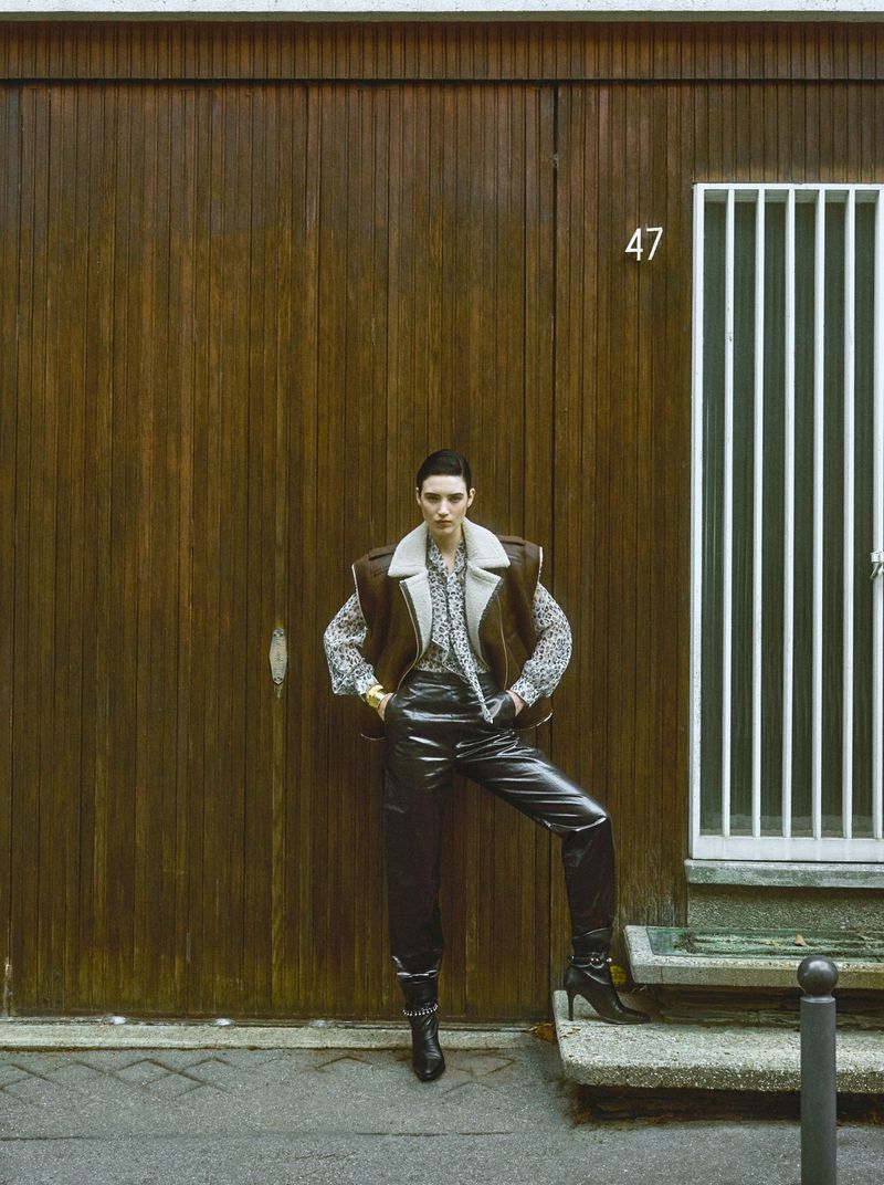 A woman in a shearling vest and leather pants stands in front of a closed wooden door, her hands on her hips in an outdoor setting.