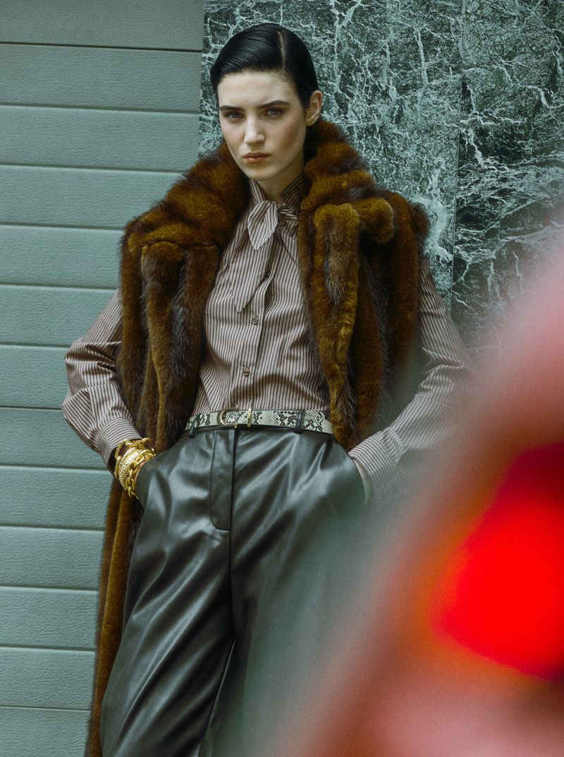 Fashionable woman in fur vest, striped shirt, and leather pants poses outdoors against an abstract marbled background.