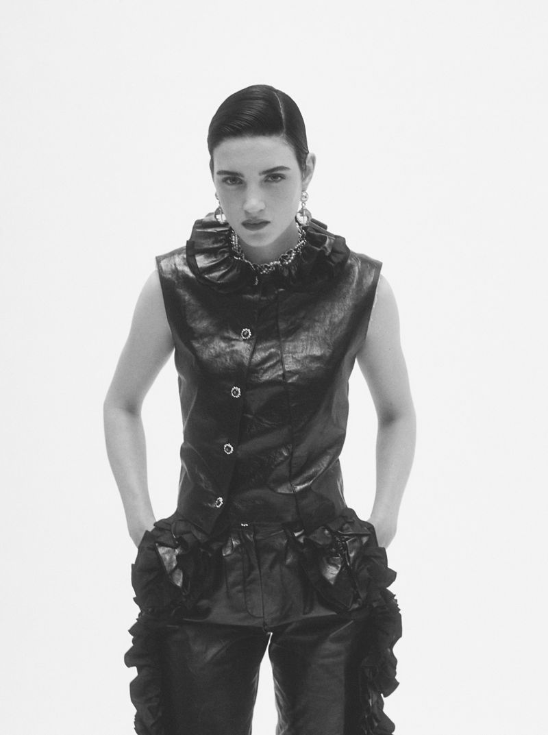 Model Eloise Logan poses in a black ruffled sleeveless leather vest and matching trousers against a plain white studio backdrop.
