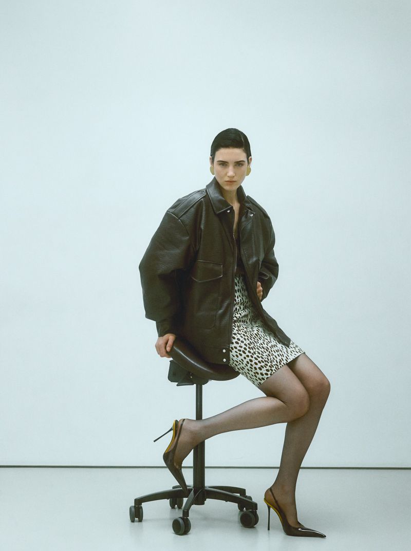 A woman poses sitting on a black rolling chair wearing a black leather jacket and animal print dress in a fashion editorial.