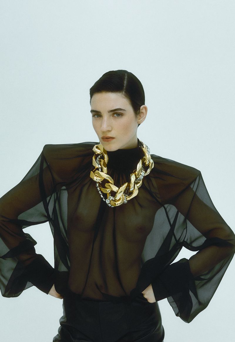 A slender woman poses indoors wearing a sheer black blouse, black pants, and a chunky gold chain necklace against a white backdrop.