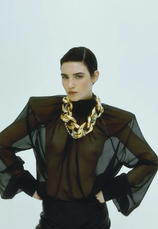 A slender woman poses indoors wearing a sheer black blouse, black pants, and a chunky gold chain necklace against a white backdrop.