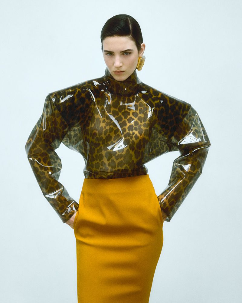 Fashion portrait of a woman with dark hair wearing a leopard-print top with a yellow skirt, standing against a white background in a studio.