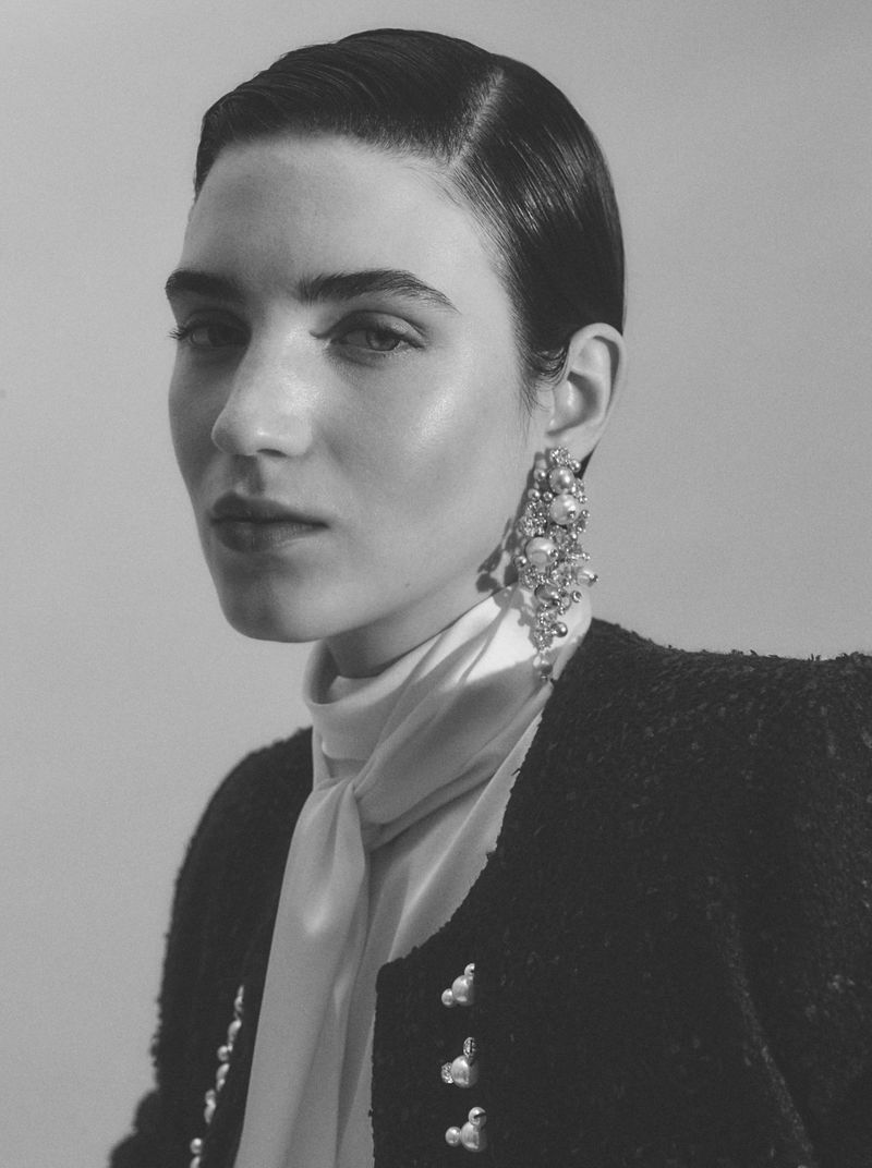 Close-up of a woman with slicked-back hair wearing statement earrings, a bouclé jacket, and a light scarf, captured in soft studio lighting.