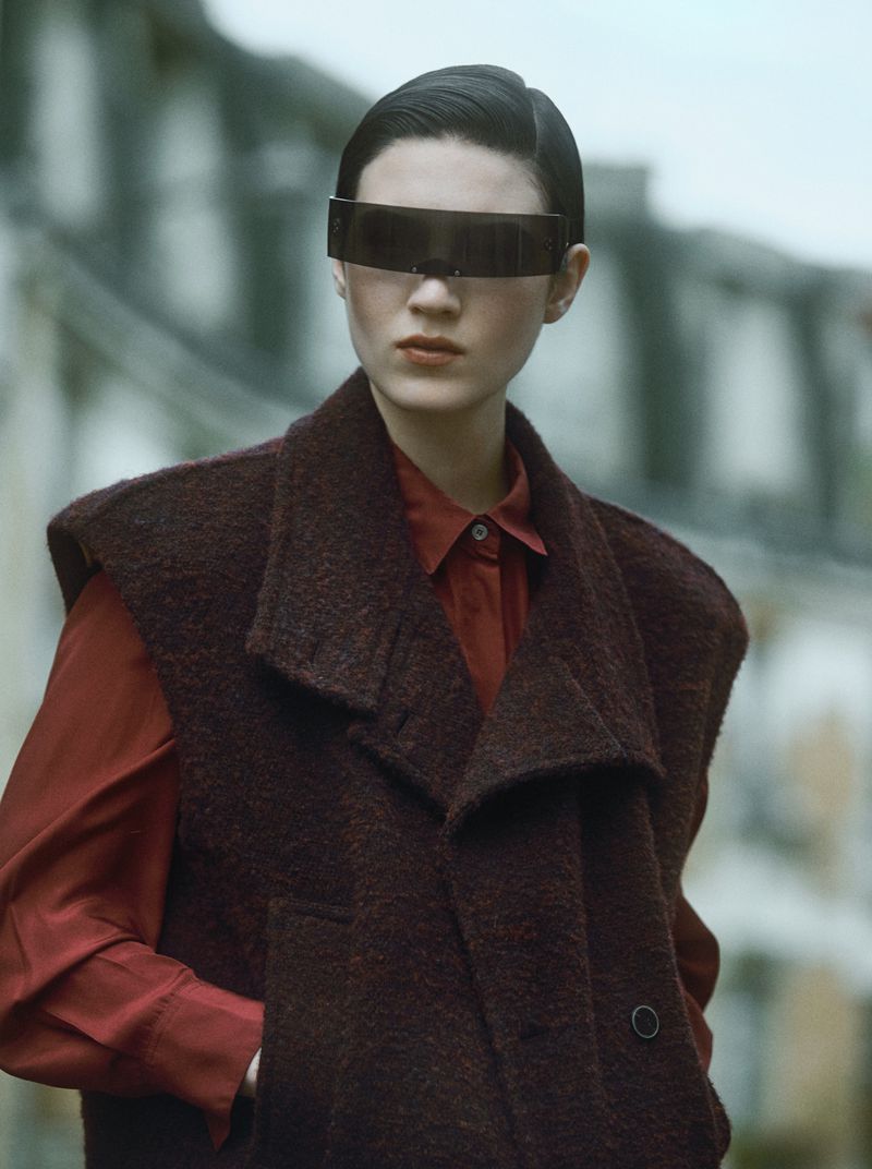 Elegant woman with futuristic visor-like sunglasses, burgundy vest and red blouse in urban setting, styled in an editorial fashion shoot.