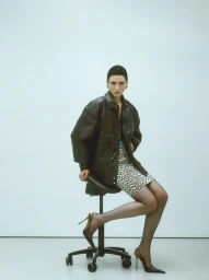 Model Eloise Logan sitting on a rolling stool wearing an oversized dark leather jacket, patterned mini dress, and slingback heels.
